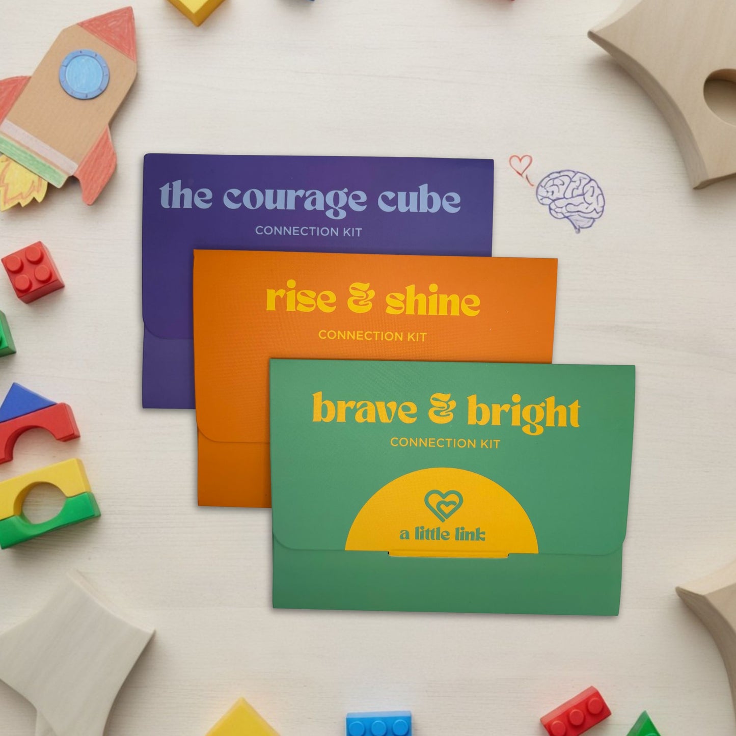 Coluorful A Little Link 'Connection Kit' envelopes on a light surface with educational toys around. This bundle includes The Courage Cube, Rise and Shine, and Brave and Bright